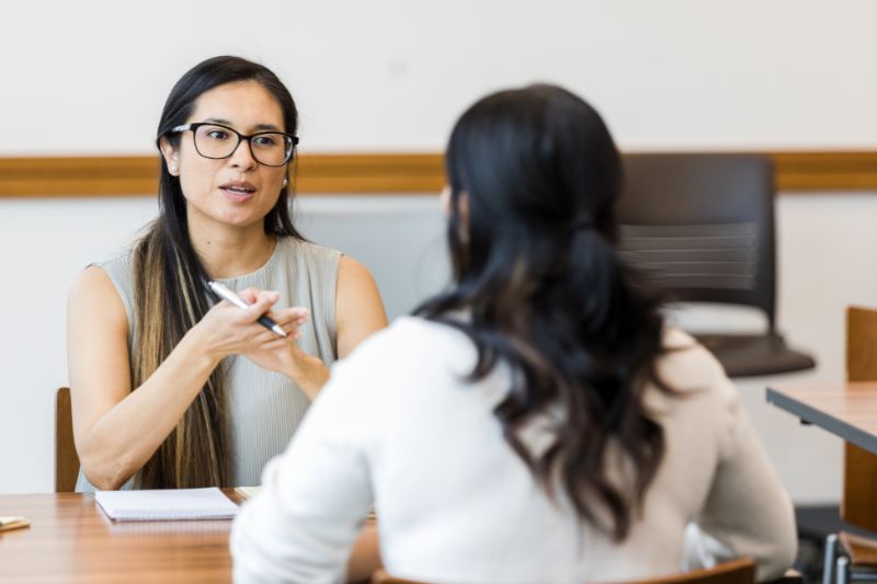 Long Dark haired lady with dark framed glasses sitting at a table with a pen and notepad talking to another dark haired female wearing and white long sleeved top.